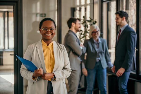 Smiling businesswoman in the office of a purpose-led company with colleagues discussing CSR in the background